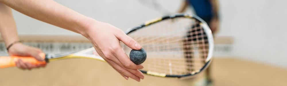 Close-up of woman's hands as she holds a squash ball up to a racquet, preparing to serve on a squash court